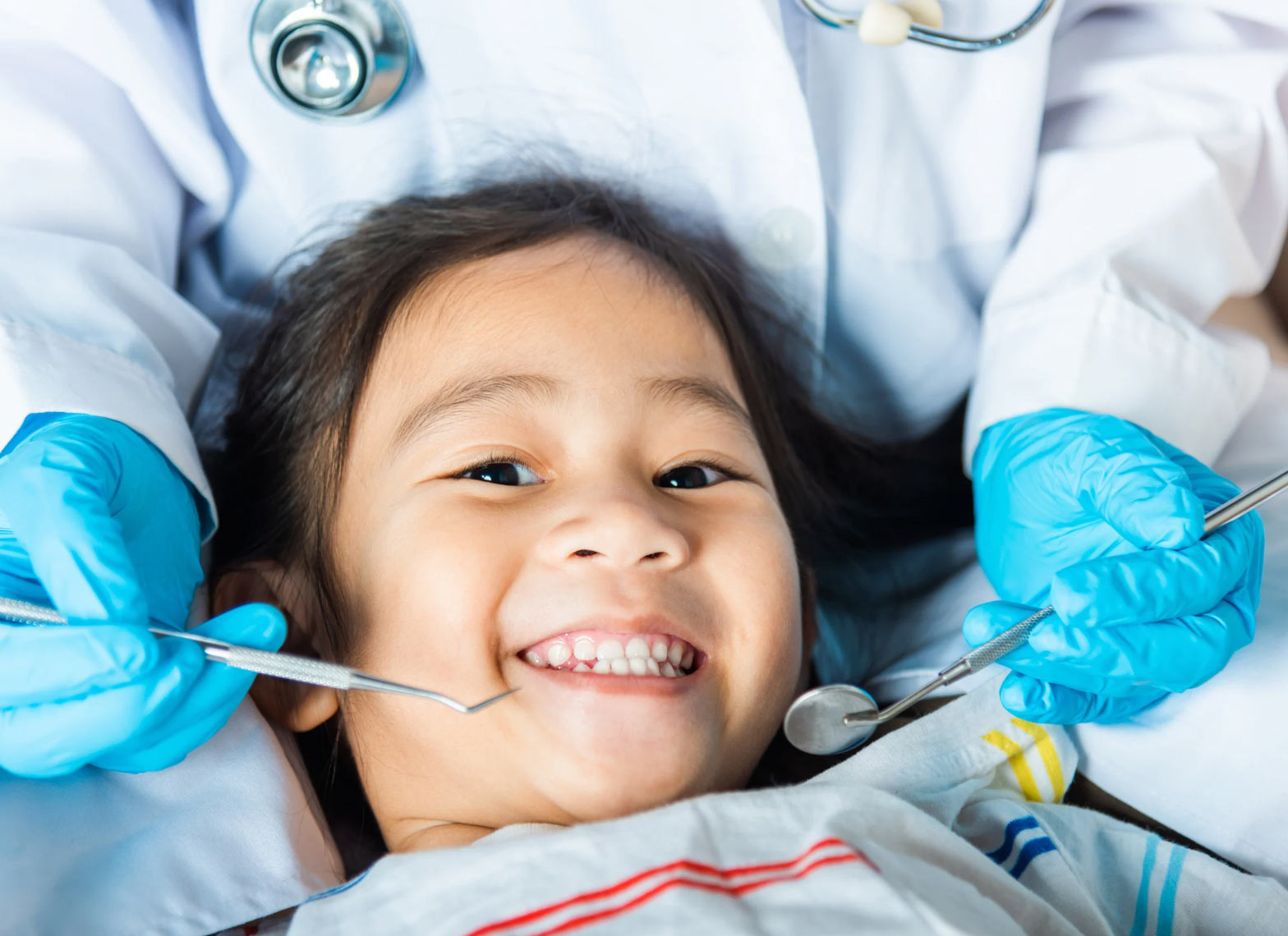 Smiling child at the dentist with blue-gloved hands holding dental tools near her mouth.
