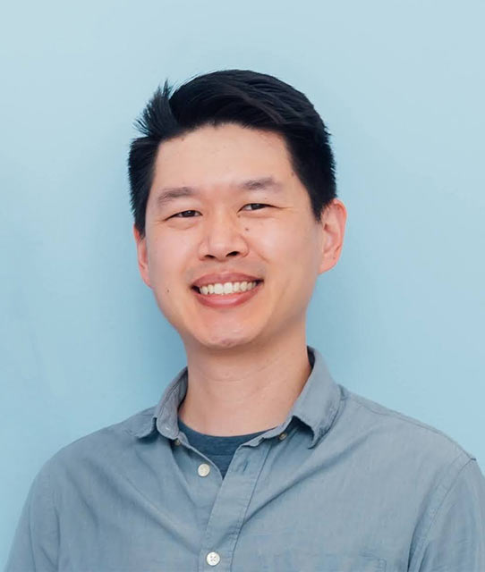 dr-chen Smiling man with short black hair in a light blue shirt standing against a pale blue background.