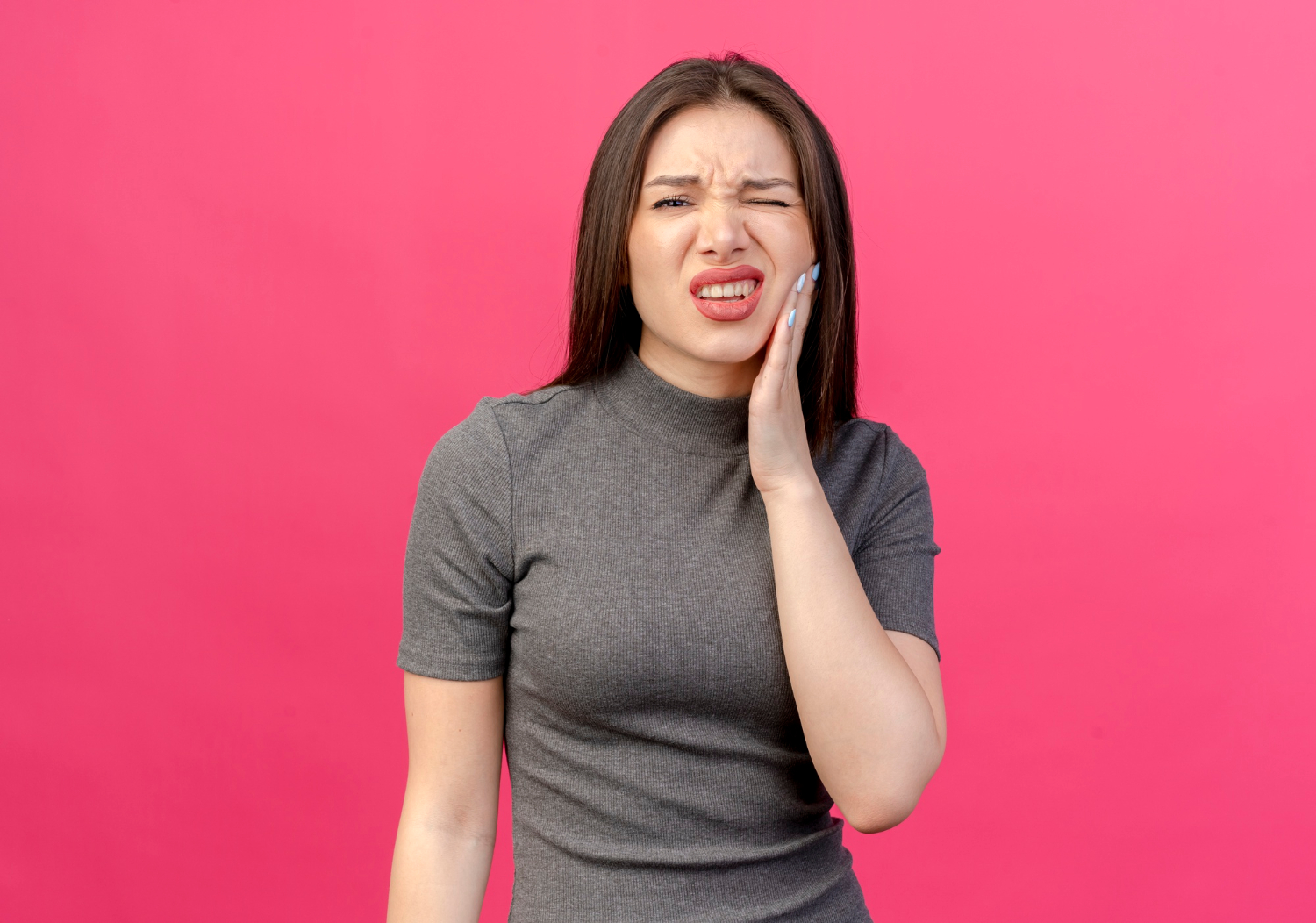 Woman with long brown hair touching her cheek and wincing in pain against a pink background.